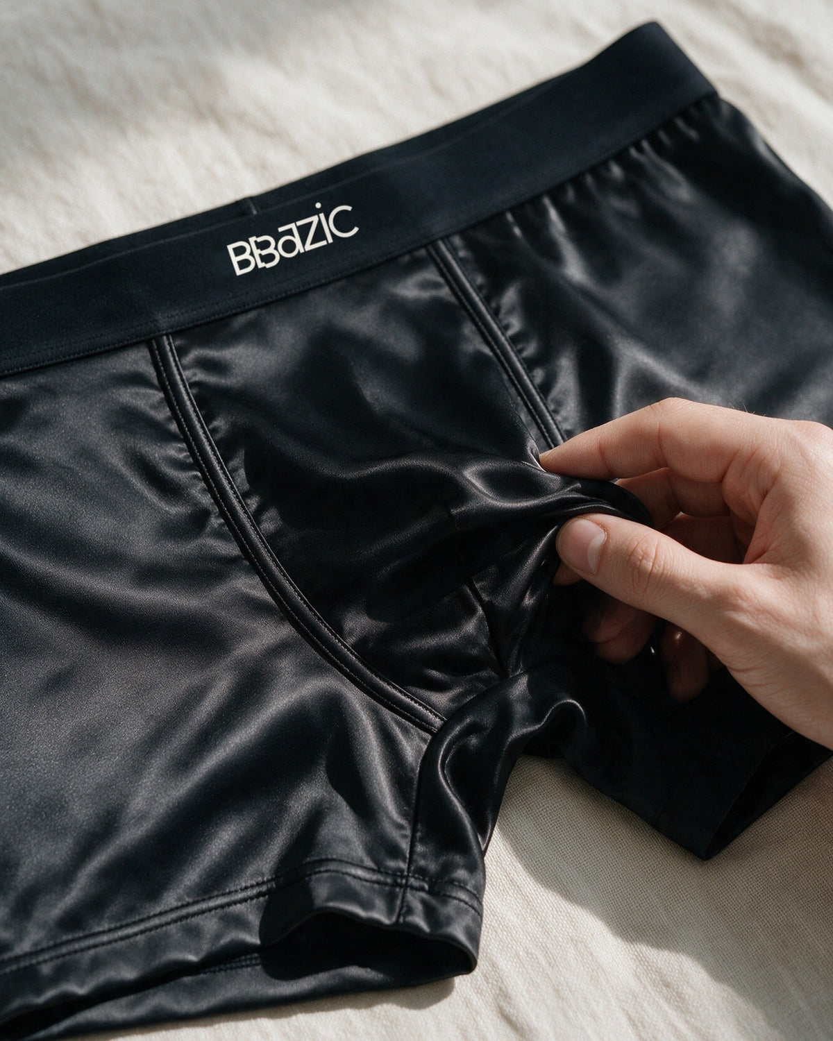 Mayfair men's silk-blend boxer in Black, fingertips lifting the silk-blend satin, BBazic logo on the waistband